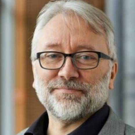 Man with gray hair and beard, wearing glasses and a suit, smiling slightly in an indoor setting at Hochschule Coburg.