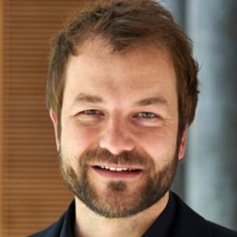 A man with short brown hair and a beard smiles warmly at the camera. He is wearing a black shirt or jacket, standing against a blurred brown and gray background, reminiscent of the academic ambiance at Hochschule Coburg.