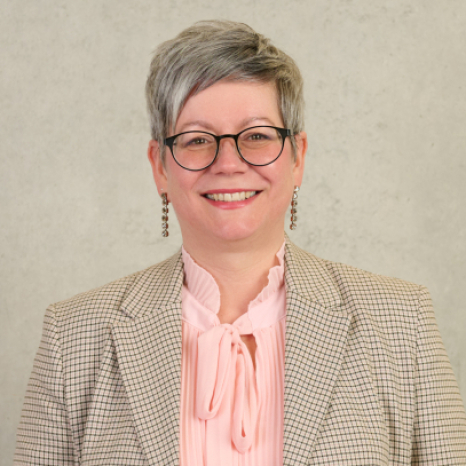 A person with short gray hair and glasses is smiling warmly, sporting a plaid blazer over a pink blouse with a bow. Their confident demeanor reflects the scholarly excellence of Hochschule Coburg. The serene backdrop is a plain light gray wall, emphasizing their sophisticated style.