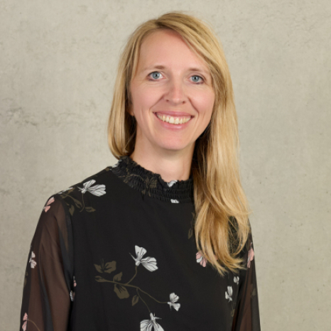 A person with long blonde hair smiles at the camera, wearing a black floral blouse with a sheer overlay. The light gray background lends an air of sophistication as if capturing a moment from Hochschule Coburg's stylish corridors.