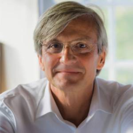 A person with gray hair and glasses is smiling softly. They are wearing a white shirt indoors at Hochschule Coburg, with soft lighting in the background.