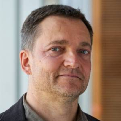 A person with short dark hair and a beard, wearing a dark blazer over a light shirt, is looking at the camera. The setting hints at Hochschule Coburg, with its blurred indoor background and soft lighting creating an academic ambiance.