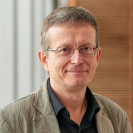 A man with short hair and glasses, possibly an instructor from Hochschule Coburg, is wearing a gray blazer over a black shirt. He is smiling slightly while standing indoors, against a blurred background.