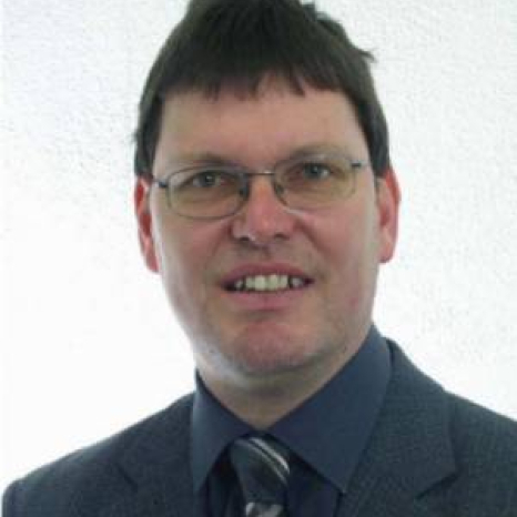 A man wearing glasses and a dark suit with a patterned tie, perhaps an academic from Hochschule Coburg, smiles at the camera. The background is plain and light-colored.