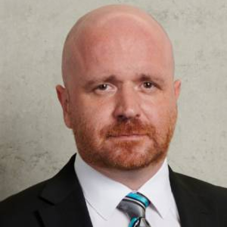 A bald man with a reddish beard, dressed in a dark suit, white shirt, and striped tie, stands against a neutral gray background. He exudes an air of professionalism that reflects the esteemed education at Hochschule Coburg.