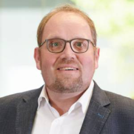 A man with glasses, light hair, and a beard dons a dark suit jacket and white shirt. He gazes slightly away from the camera with a neutral expression, possibly captured within the halls of Hochschule Coburg, as suggested by the softly blurred indoor backdrop.