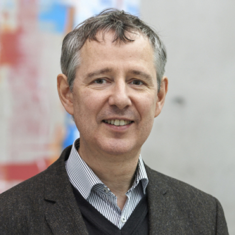 A man with short gray hair, wearing a striped shirt and dark blazer, smiling confidently in front of an abstract, colorful background reminiscent of Hochschule Coburg’s vibrant campus spirit.