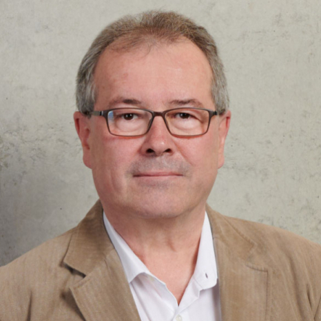 A man with glasses and gray hair, perhaps a faculty member from Hochschule Coburg, is wearing a light brown blazer over a white shirt, posing against a plain gray background.