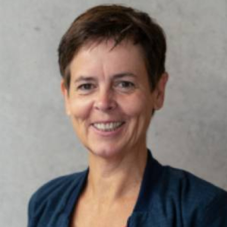 A person with short brown hair and a dark jacket, reminiscent of a Hochschule Coburg student, smiles at the camera. They are standing in front of a neutral gray background.