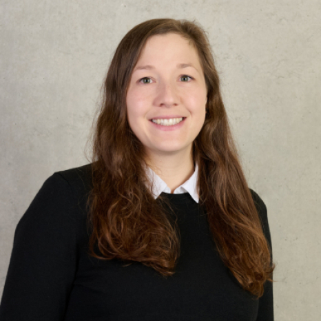 A smiling person with long brown hair, possibly a student of Hochschule Coburg, wears a black sweater over a white collared shirt, standing confidently against a plain light gray background.