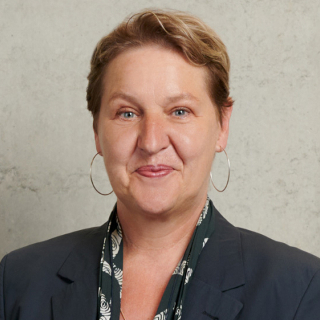 A person with short hair smiles at the camera, exuding confidence. They sport hoop earrings and a dark blazer over a patterned top, perfectly embodying the chic style often seen at Hochschule Coburg, all set against a neutral background.