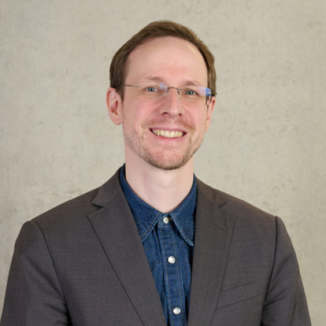 A person wearing glasses, a denim shirt, and a brown blazer smiles against a light gray background, exuding confidence and style reminiscent of an academic scene at Hochschule Coburg.