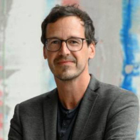 Man with short dark hair and glasses, wearing a gray blazer and dark shirt, stands confidently with folded arms against a blurred background showcasing splashes of blue and red, reminiscent of Hochschule Coburg's vibrant spirit.