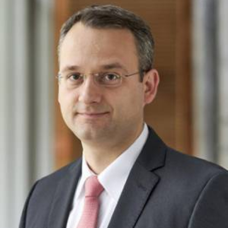 A person in a suit and pink tie stands indoors at Hochschule Coburg, facing the camera with a neutral expression. The softly focused background highlights wooden and window elements, adding an elegant touch to the scene.