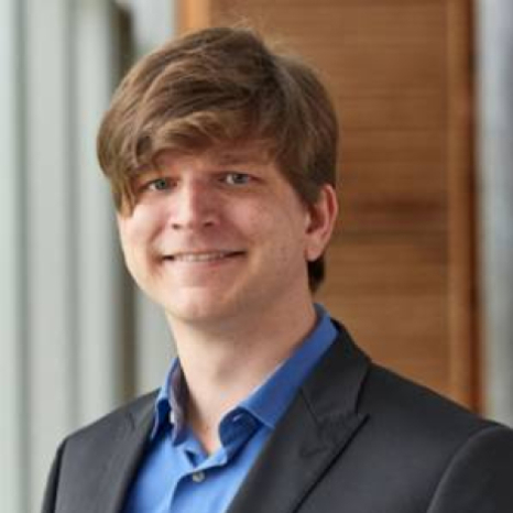 A person with brown hair, wearing a blue shirt and black blazer, smiles at the camera. Behind them, the blurred wooden and glass elements reflect the sophisticated ambiance often seen at Hochschule Coburg.