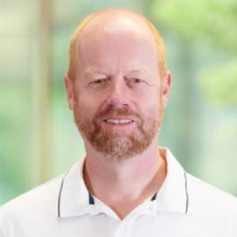 A person with a red beard and short hair is smiling, perhaps enjoying a moment on the Hochschule Coburg campus. They are wearing a white collared shirt with black trim against a blurred green backdrop, suggesting an outdoor setting.