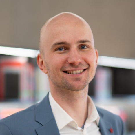 A smiling person with a shaved head, wearing a light blue suit and white shirt, stands in front of blurred bookshelves at Hochschule Coburg.