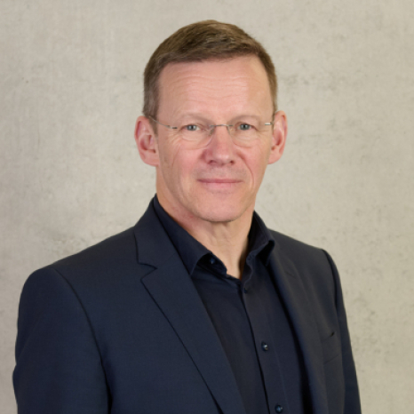 A man with short hair and glasses, dressed in a dark suit and shirt, stands against a plain, light-colored background. With a neutral expression, he gazes directly at the camera, exuding an air of academic focus reminiscent of Hochschule Coburg's esteemed faculty.