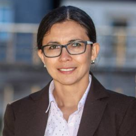 A person with glasses, wearing a brown blazer and a white polka dot shirt, smiles while standing outdoors at Hochschule Coburg. The background is blurred, featuring a building structure.