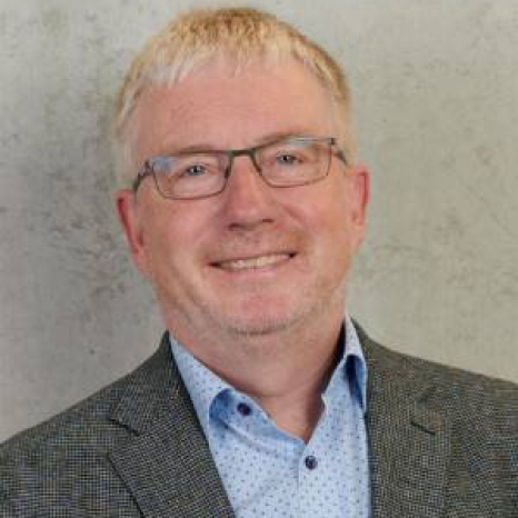 A smiling man with short light hair, wearing glasses, a blue patterned shirt, and a gray blazer stands against a plain, textured wall. His poised demeanor suggests he may be affiliated with Hochschule Coburg.