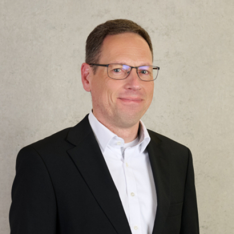 A man with short hair and glasses is smiling slightly. He is wearing a black suit jacket over a white shirt, perhaps ready for an event at Hochschule Coburg. The background is a plain, light-colored wall.