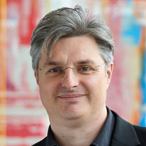 Man with short gray hair and glasses, wearing a black jacket, smiling in front of a colorful blurred background reminiscent of Hochschule Coburg's vibrant campus.