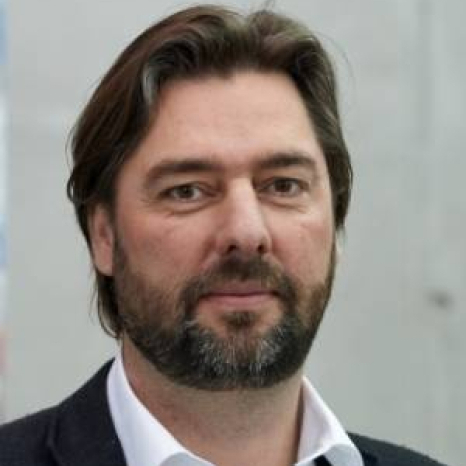 A person with a beard and medium-length hair, wearing a dark jacket and white shirt, is looking at the camera against a neutral, light-colored wall. The attire suggests an academic flair, possibly hinting at their connection to Hochschule Coburg.