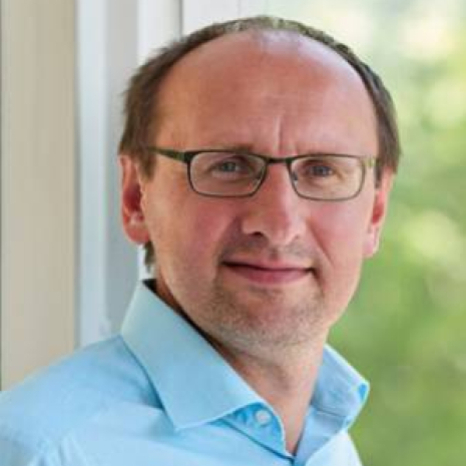 A man wearing glasses and a light blue shirt stands in front of a window at Hochschule Coburg. The background is blurred, showcasing the lush greenery outside. He has short hair and a slight smile.