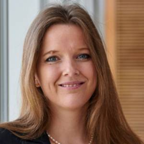 A woman with long brown hair and blue eyes smiles at the camera, embodying the sophistication of Hochschule Coburg. She wears a black top paired with a pearl necklace, set against a backdrop of vertical window blinds and wooden panels.