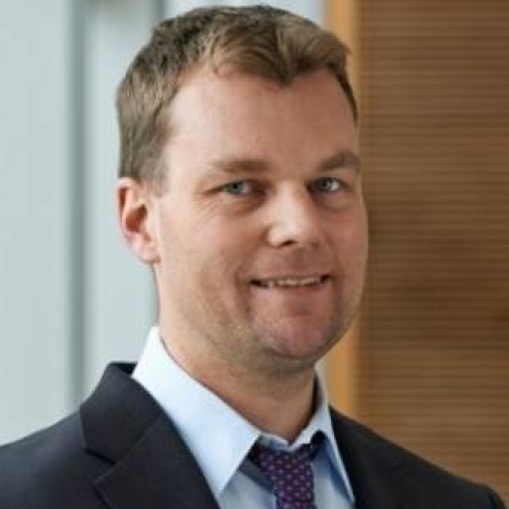 A man wearing a black suit, light blue shirt, and purple tie is smiling slightly against a blurred interior background with wooden paneling and a window. It's as if he's at Hochschule Coburg, blending seamlessly into its refined yet inviting atmosphere.