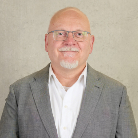 A bald man with glasses and a goatee, representing Hochschule Coburg, smiles at the camera in a grey suit jacket over a white shirt. He stands against a plain, light-colored background.