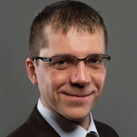 A man wearing glasses, a dark suit, and a light shirt is smiling against a plain gray background, exuding an air of confidence typical of a Hochschule Coburg alumnus.