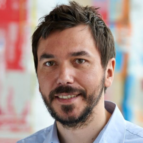 A man with short brown hair and a beard is smiling in his Hochschule Coburg light blue collared shirt. The background is blurred with various colors.