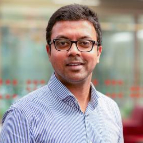 A person wearing glasses and a striped shirt smiles at the camera, exuding a friendly demeanor. The background, reminiscent of Hochschule Coburg's vibrant campus, is slightly blurred with hints of red and green hues.