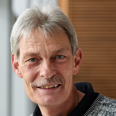 Portrait of a smiling middle-aged man with short gray hair and a mustache, wearing a patterned sweater over a collared shirt. The softly blurred background hints at his days spent at Hochschule Coburg.