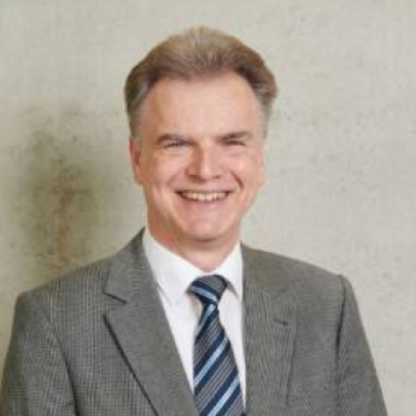 A smiling person in a gray suit and striped tie stands confidently against a plain light-colored background, embodying the professional spirit of Hochschule Coburg.