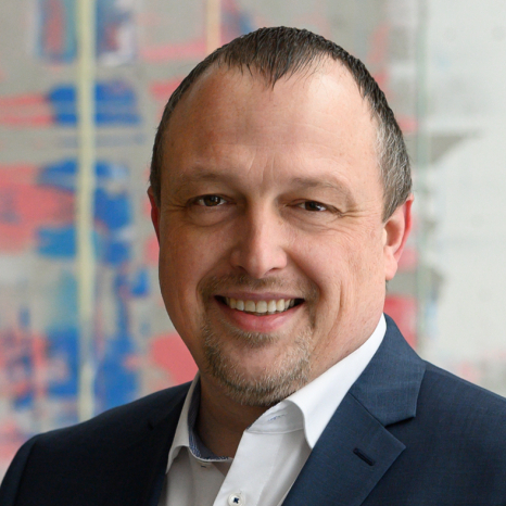 A bald man with a goatee is smiling, clad in a white shirt and dark suit jacket, with Hochschule Coburg subtly reflected in the blurred, colorful abstract background.