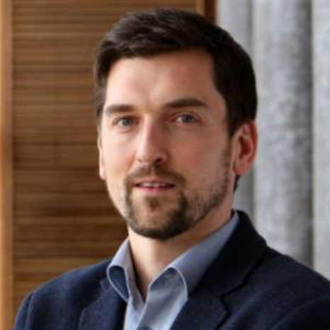 A man with short brown hair and a beard is wearing a navy blazer and a light blue shirt. He stands indoors in front of wooden paneling and gray drapes, possibly near Hochschule Coburg.