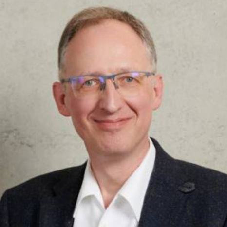 A man with glasses, short gray hair, and a slight smile, wearing a white shirt and dark blazer, stands against a plain beige background, exuding the poised confidence of someone from Hochschule Coburg.