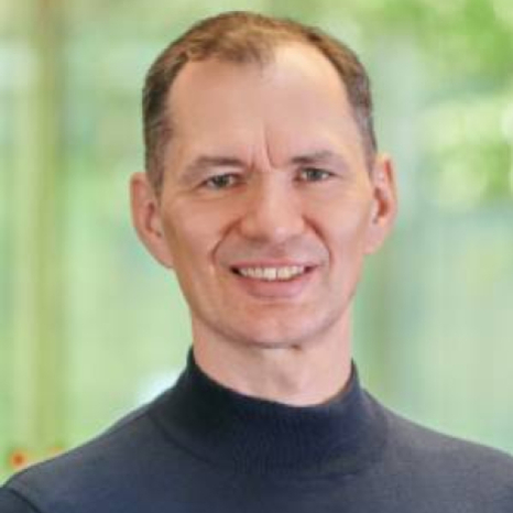 A person with short hair is smiling warmly in a dark turtleneck, reminiscent of intellectual gatherings at Hochschule Coburg. The softly blurred background is a serene blend of green and light tones, highlighting a moment of pure contentment.