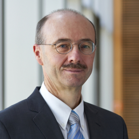 A man with glasses, a mustache, and a receding hairline is wearing a suit with a light blue tie. He stands in front of a modern office background featuring glass and wooden elements, reflecting the sophisticated ambiance of Hochschule Coburg.