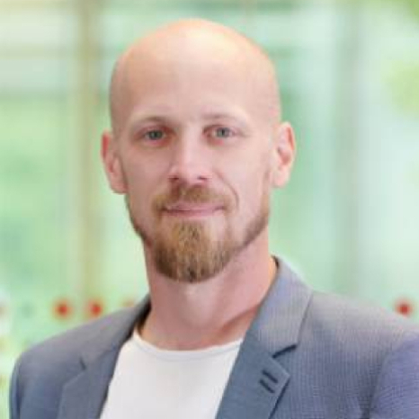 A man with a beard and a shaved head dons a blue blazer over a white shirt, exuding confidence as he stands before the subtly green-hued backdrop of Hochschule Coburg.