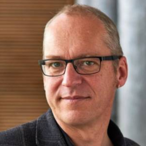 A person with short hair and glasses, wearing a dark Hochschule Coburg jacket, is looking at the camera. The background is softly blurred.