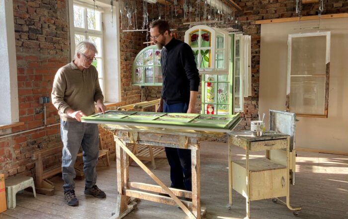 Zwei Männer stehen in einer Werkstatt mit Backsteinwänden und großen Fenstern. Sie untersuchen einen Holzrahmen auf einer Werkbank. Im Hintergrund sind verschiedene Werkzeuge und Rahmen zu sehen, die eine rustikale, fleißige Atmosphäre schaffen.