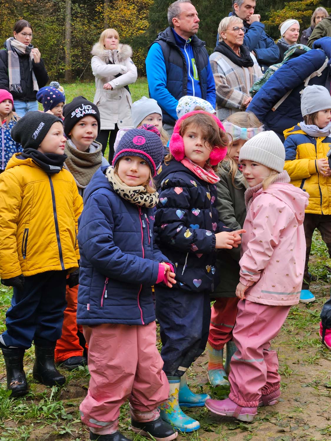 Eine Gruppe kleiner Kinder, dick eingepackt in Winterkleidung, steht draußen zusammen, manche tragen bunte Jacken, Mützen und Gummistiefel. Um sie herum stehen Erwachsene und beobachten. Die Szene scheint ein Park oder ein Waldgebiet zu sein.