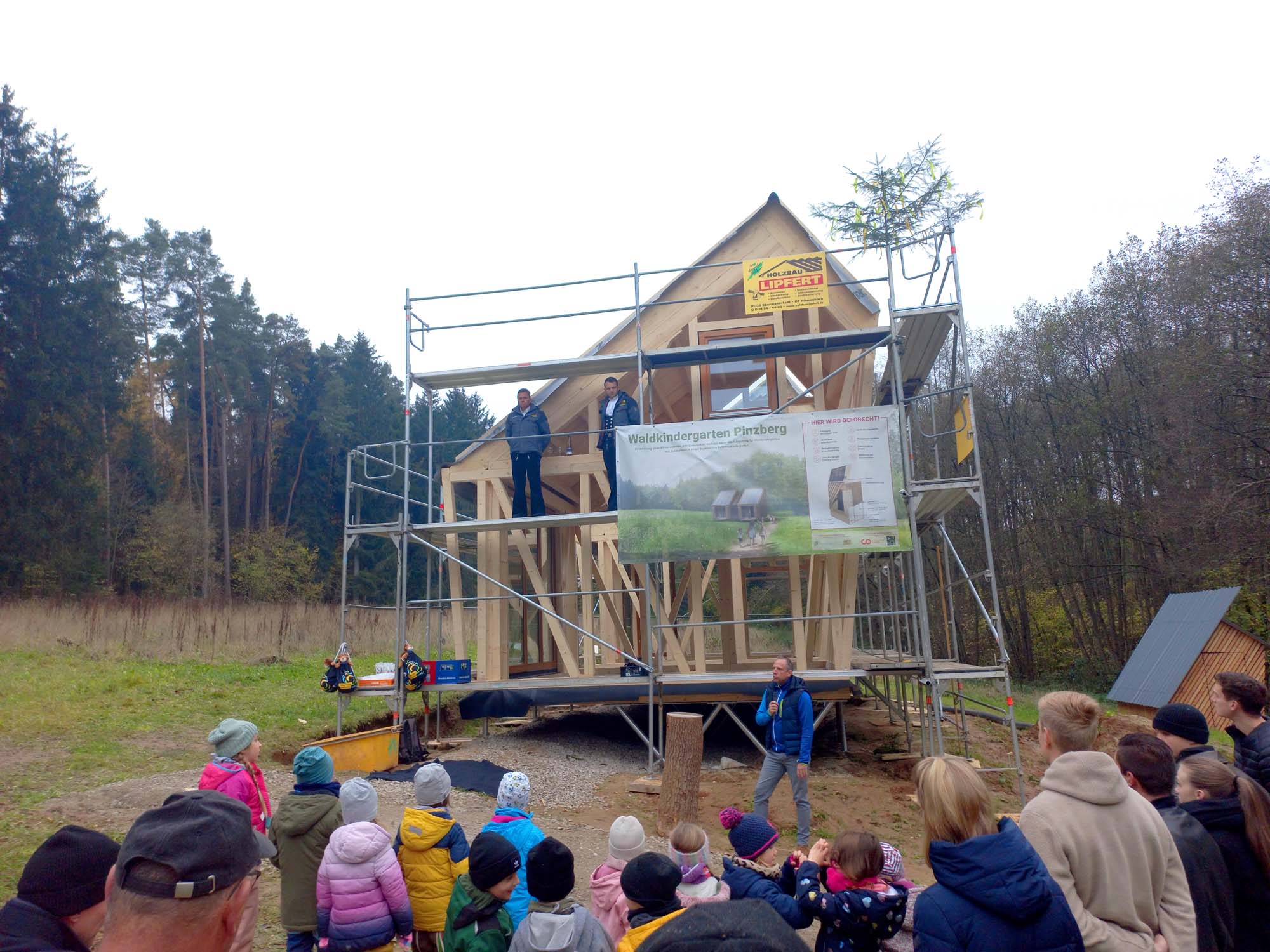 Eine Gruppe von Menschen, darunter Kinder in bunten Jacken, versammelt sich um eine im Bau befindliche Holzkonstruktion in einem Waldgebiet. Arbeiter stehen auf Gerüsten und an der Konstruktion hängen Banner.