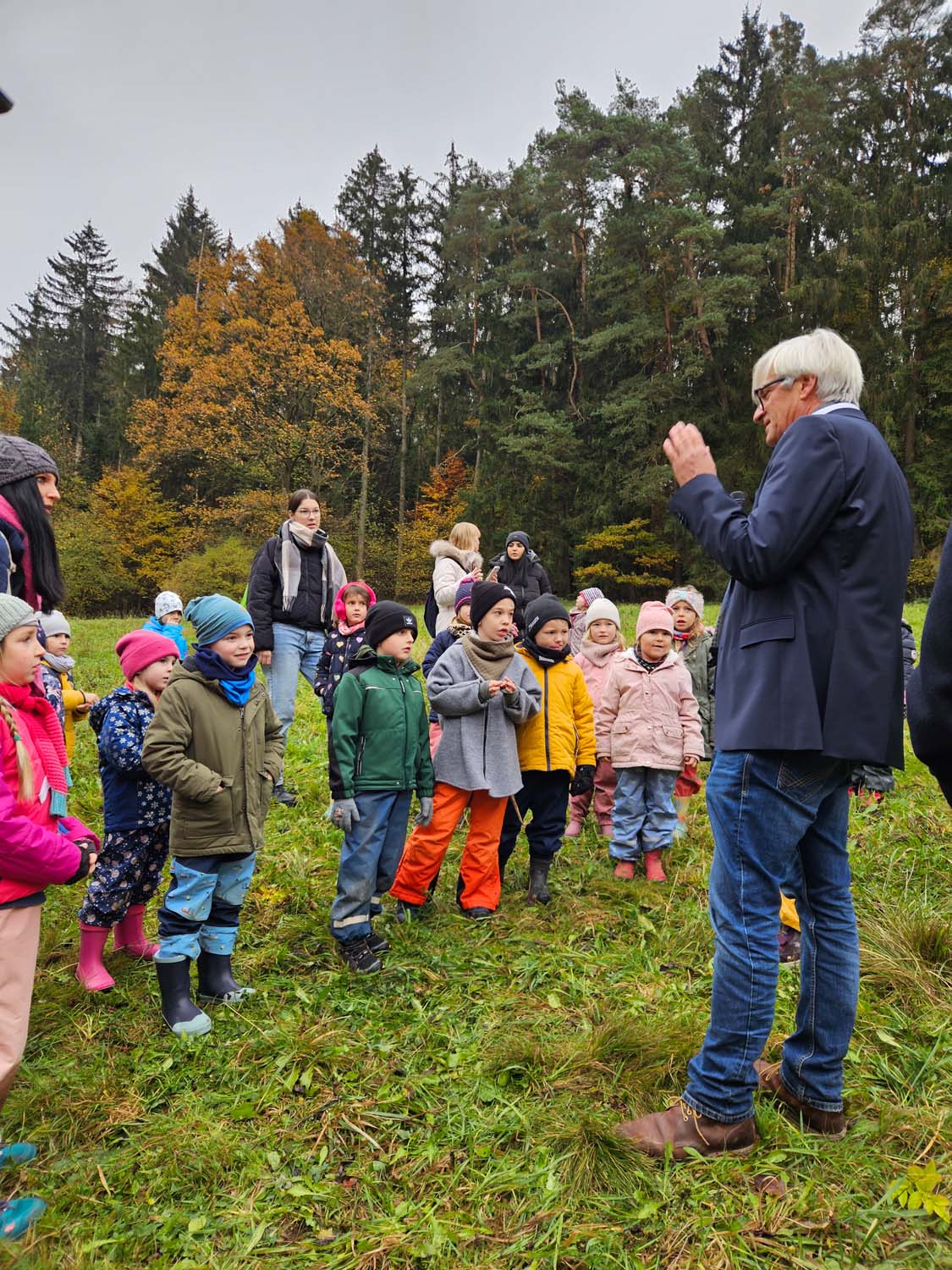 Eine Gruppe von Kindern und Erwachsenen versammelt sich um einen älteren Mann, der auf einer von Bäumen umgebenen Rasenfläche spricht. Die Kinder tragen farbenfrohe Outdoor-Kleidung, die für kühles Wetter geeignet ist. Die Szene vermittelt eine lehrreiche oder naturbezogene Outdoor-Aktivität.