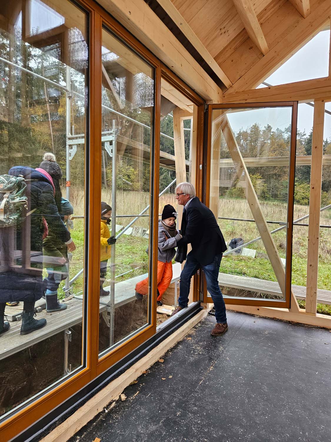 Ein Mann im Anzug mit Schal lehnt im Inneren eines Holzgebäudes an einer Glastür und interagiert mit einer Person draußen, die Winterkleidung trägt. Im Hintergrund sind weitere Personen, ebenfalls in Winterkleidung, auf einem Gerüstpodest zu sehen.