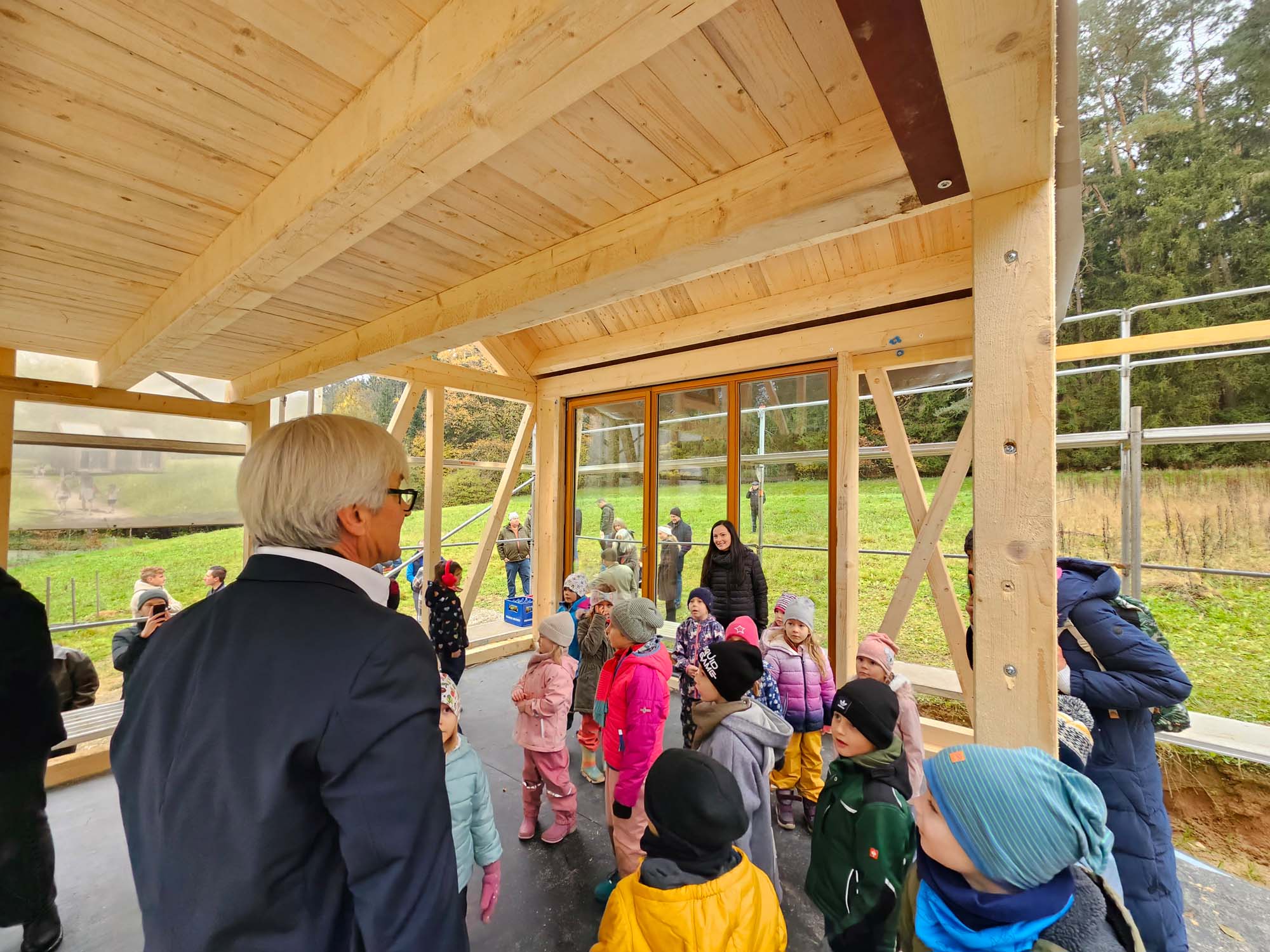 Eine Person im Anzug spricht in einem Holzbau mit einer Gruppe von Kindern, die bunte Jacken und Hüte tragen. Im Hintergrund sind Bäume und Gras zu sehen.