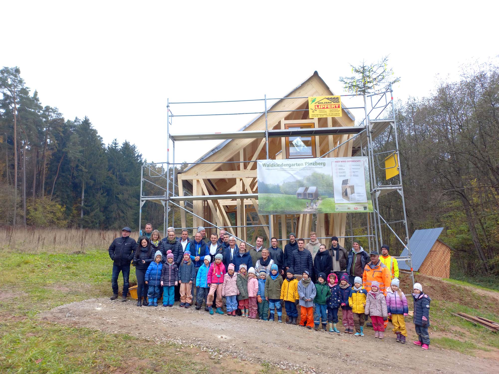 Eine Gruppe von Erwachsenen und Kindern posiert vor einem im Bau befindlichen Holzgebäude. Das Gebäude ist von einem Gerüst umgeben und liegt in einem Waldgebiet. Die Kinder tragen farbenfrohe Kleidung. Auf dem Gerüst hängt ein Banner.